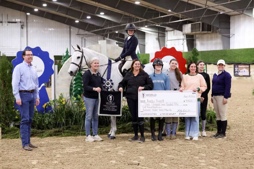A group of people pose indoors with a horse and rider; one person holds a large check and another holds an award sign. Large ribbons are displayed in the background.
