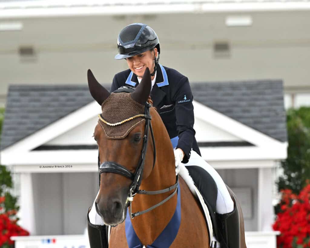 A rider in formal equestrian attire sits on a brown horse with a blue ribbon on its neck, smiling in an outdoor competition setting.