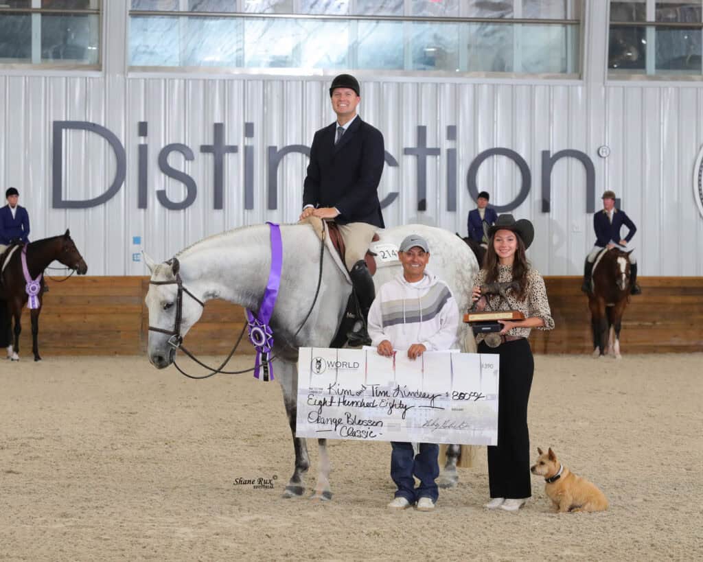 A rider on a gray horse holds a purple ribbon while two people in front display a large check and a trophy; a dog sits nearby in an indoor arena.