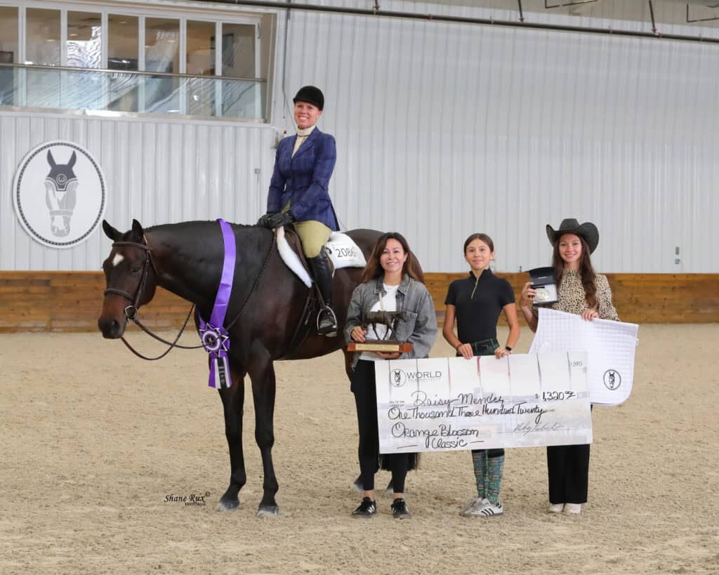 Four women pose with a horse in an indoor arena. One woman is mounted, while the others hold a trophy, a ribbon, and an oversized check for $1,350.