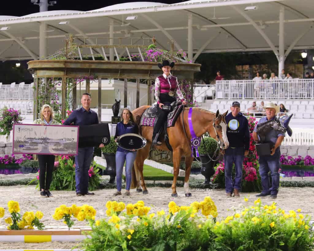 A group of people poses with awards and a horse rider in western attire on horseback, surrounded by flowers in an arena setting.