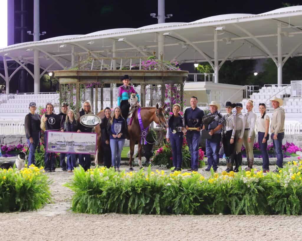 A group of people and a horse pose at an outdoor award ceremony at night, with trophies, ribbons, a large check, and greenery in the foreground.