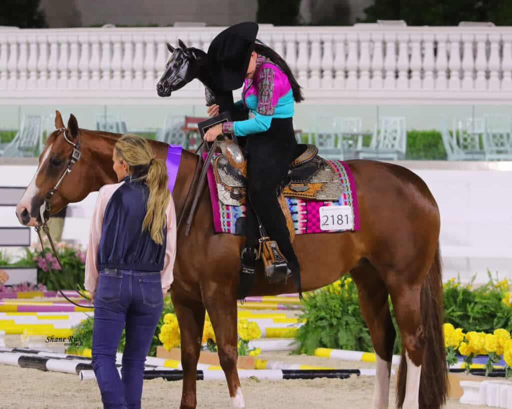 A rider in colorful attire sits on a brown horse wearing a patterned saddle blanket, holding a trophy. Another person stands nearby, both in an indoor arena with flowers and white fencing.