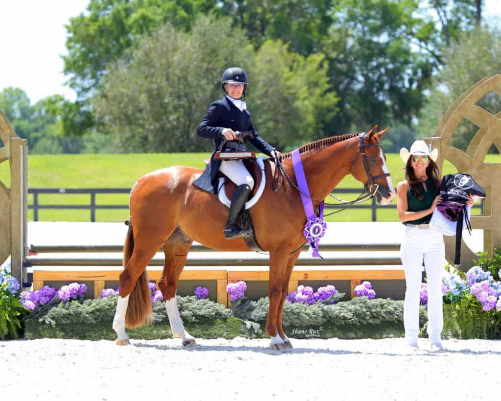 A person in equestrian attire sits on a horse with a purple ribbon, next to a standing person in a cowboy hat holding awards, at an outdoor event with flowers and wooden structures.