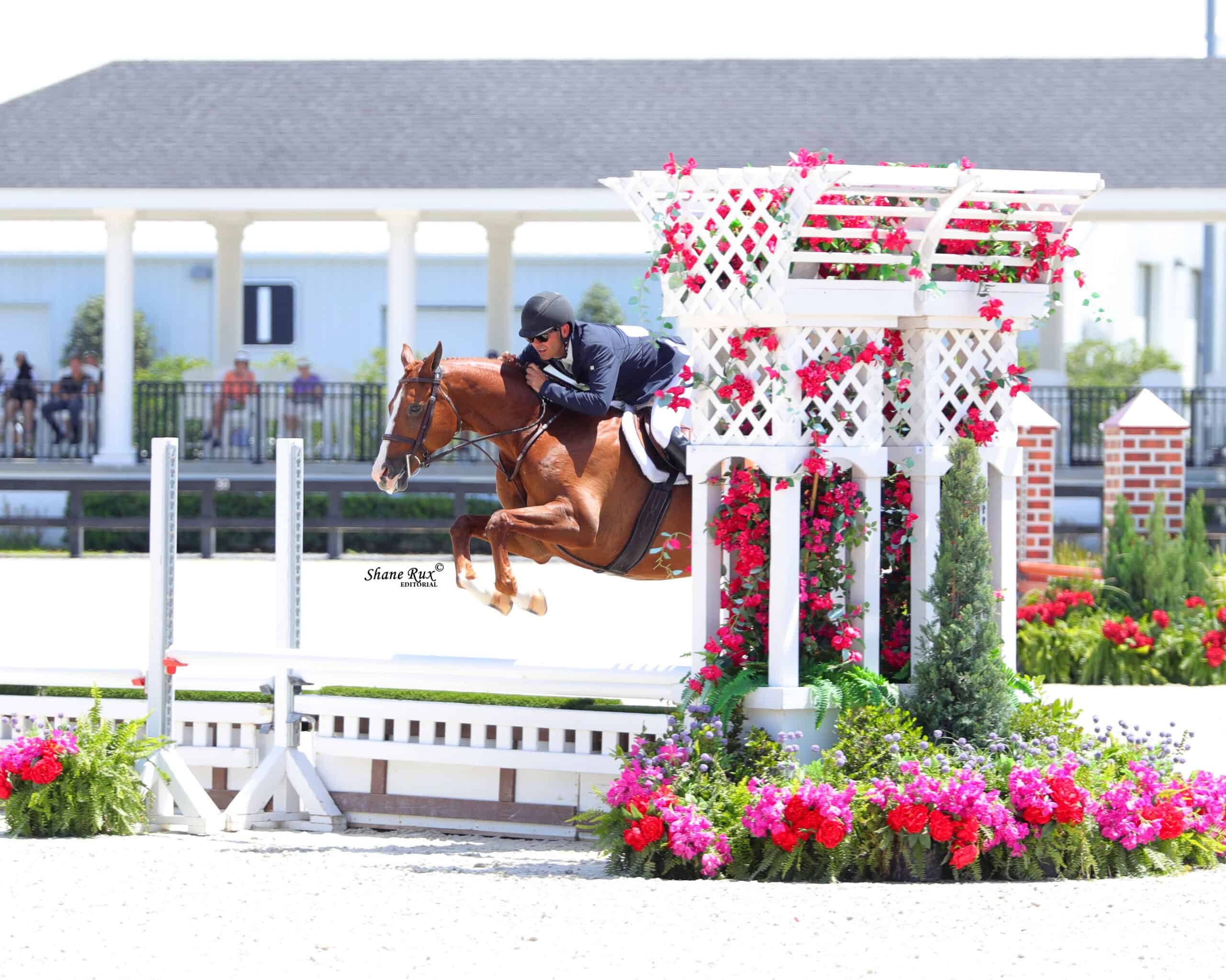 A rider on a brown horse jumps over a white fence decorated with flowers during an equestrian competition in an outdoor arena.