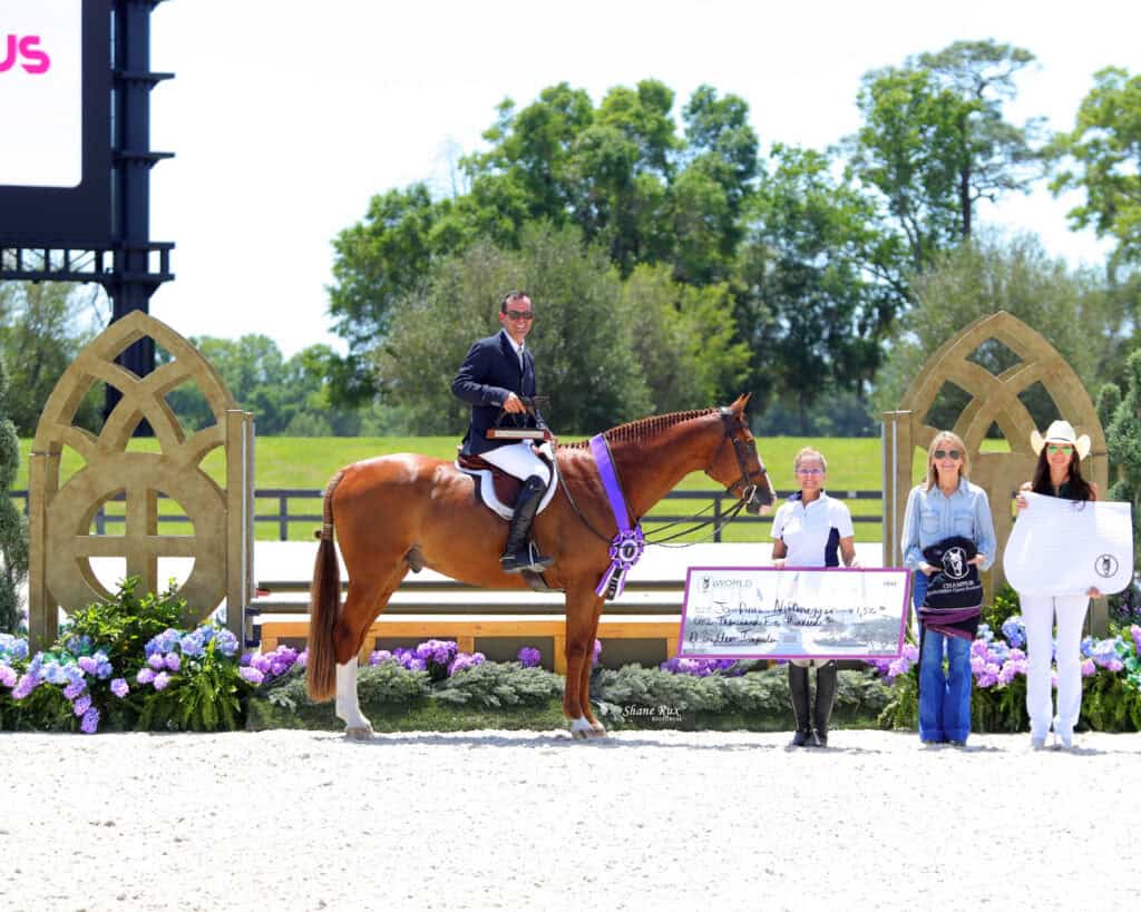 A man on horseback with a purple ribbon poses next to three women, one holding a large check, in an outdoor equestrian event setting with floral decorations.