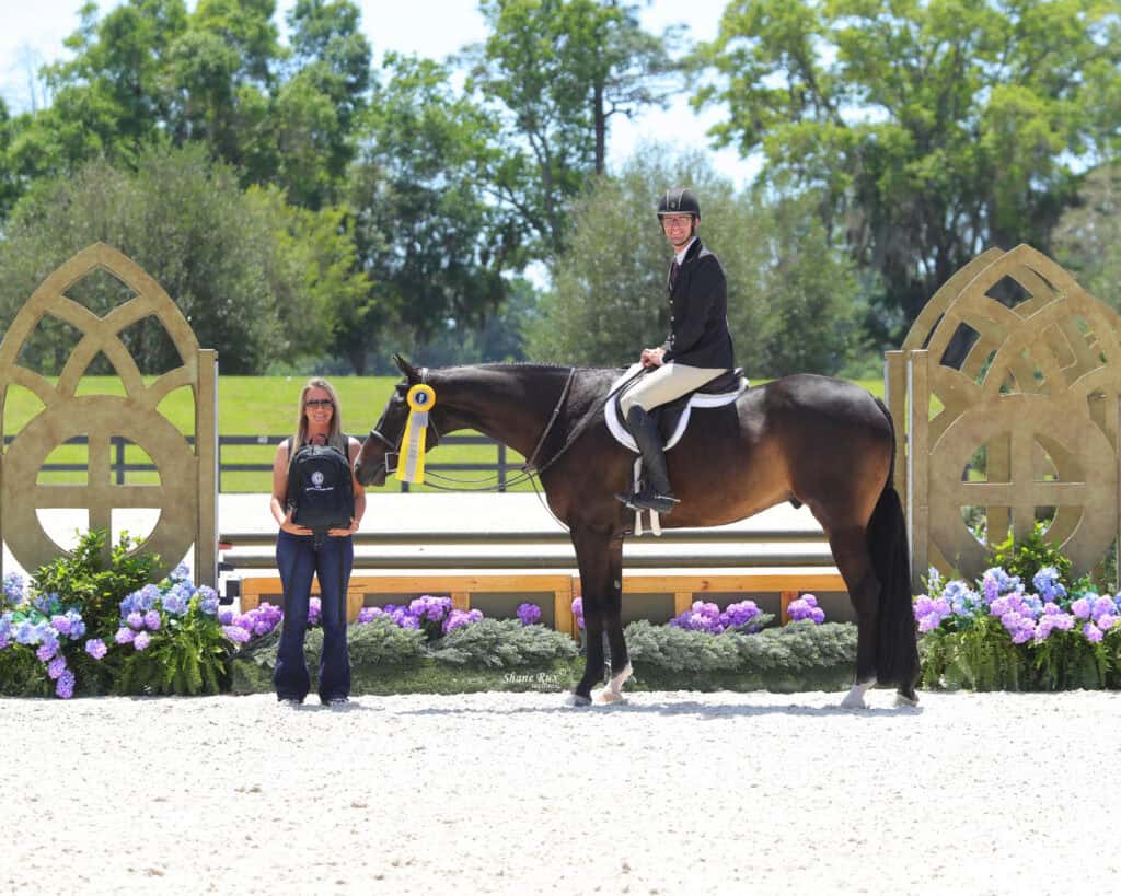 A person riding a horse with a yellow ribbon poses next to a standing woman holding a trophy, all in front of decorative wooden arches and colorful flowers.