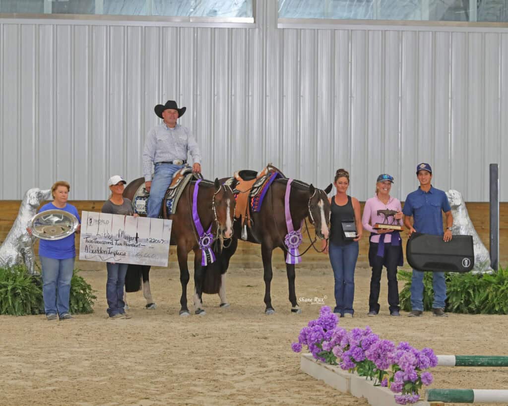 A group of six people and two horses stand indoors. The people hold awards, a large check, a tray, and a guitar case. Purple ribbons are attached to the horses.