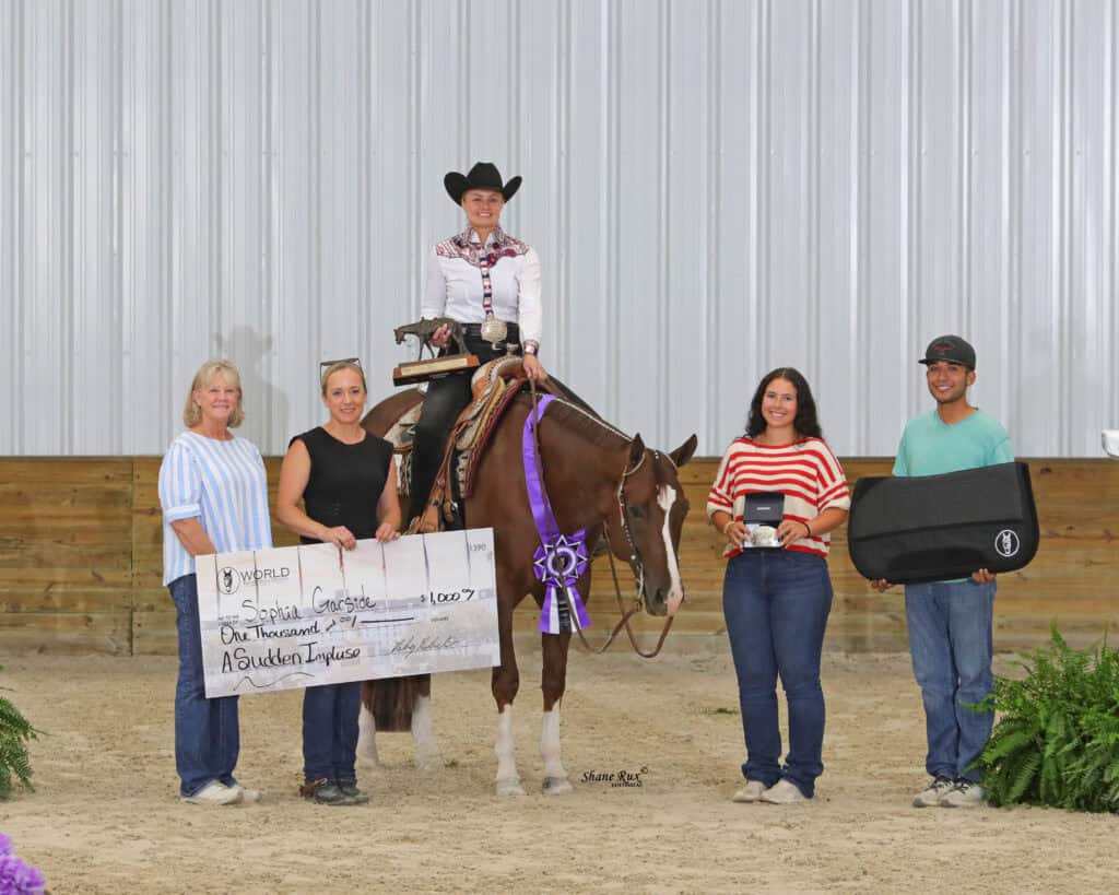 A woman on a horse holds a ribbon, surrounded by four people; another person holds a large check and another holds a prize case. They are inside a riding arena.