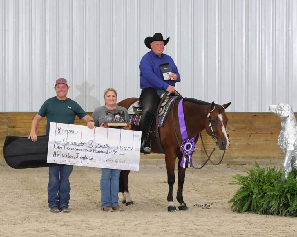 Three people pose indoors with a horse. One man sits on the horse holding a plaque, while the other two stand beside them holding a large ceremonial check and a ribbon.