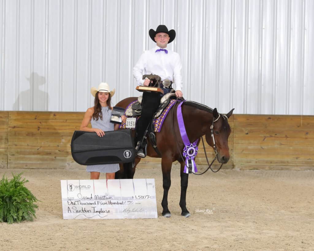 A man on horseback wearing western show attire poses with awards alongside a woman holding a large prize check and a case, both standing in an indoor arena.