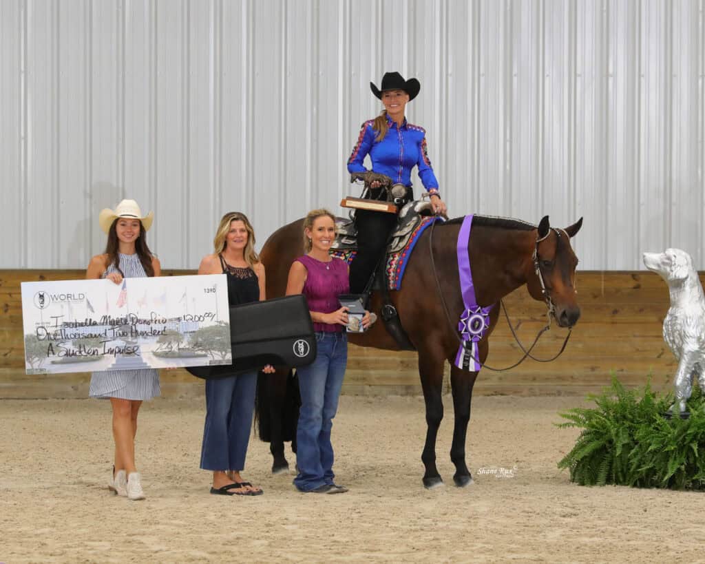 Four women pose with a horse in an indoor arena; one is mounted, another holds a large check, and there is a prize ribbon on the horse’s neck. A dog statue stands nearby.