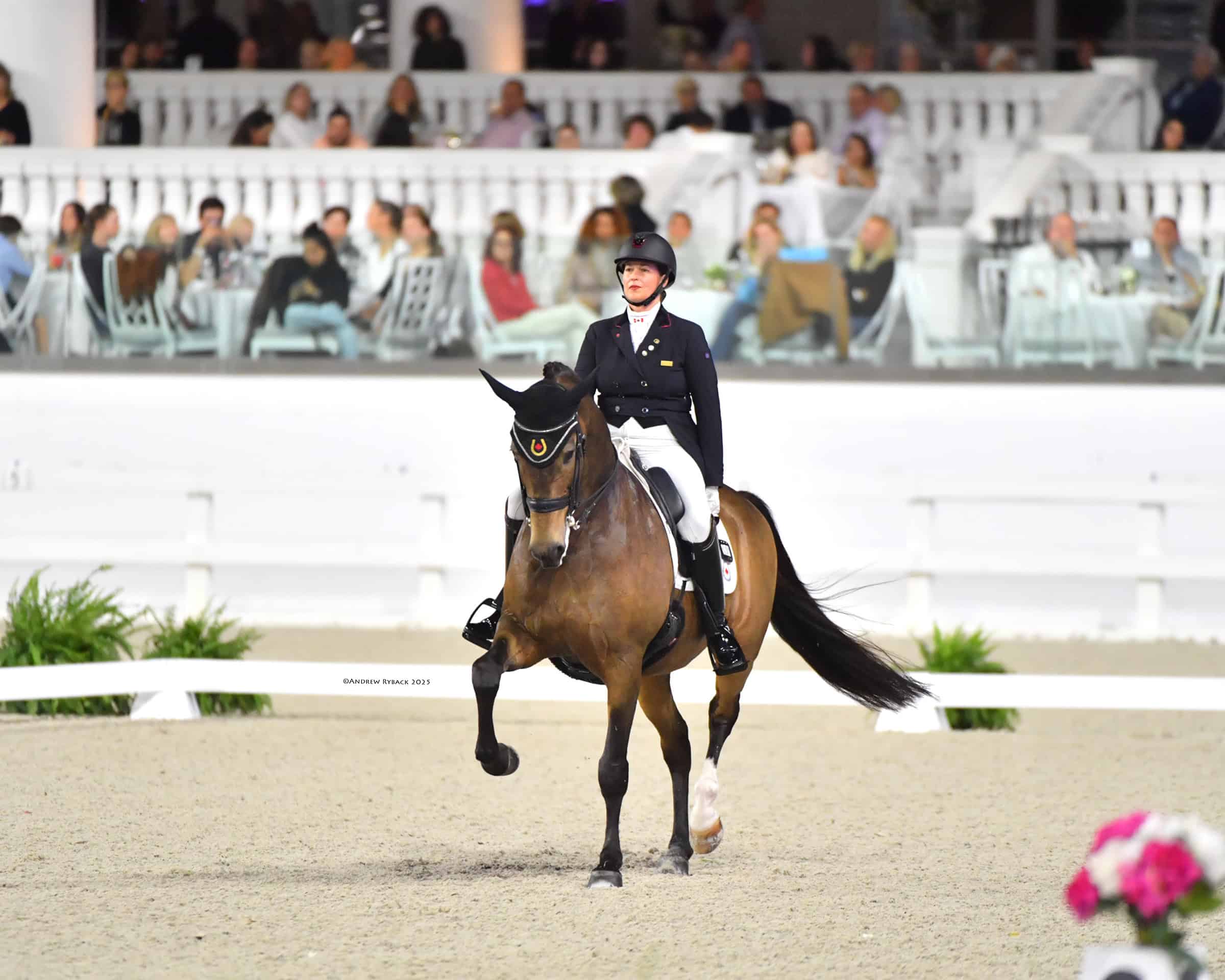 Equestrian rider in formal attire guiding a horse during a dressage competition, with spectators seated at tables in the background.