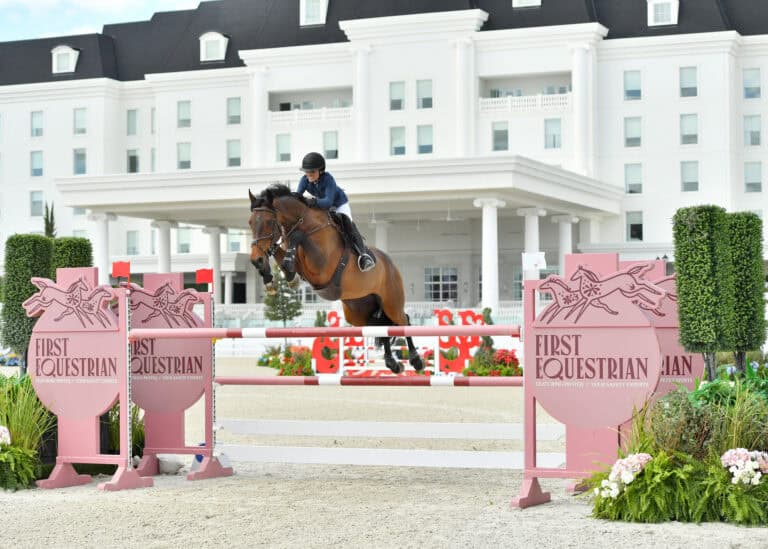 A rider on a horse jumps over an obstacle labeled "First Equestrian" at an outdoor equestrian event, with a white building in the background.