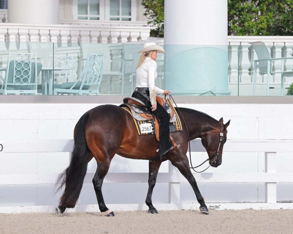 A rider in a white hat and long-sleeve shirt guides a dark brown horse along a sandy arena in front of a white building and patio furniture.