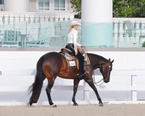 A rider in a white hat and long-sleeve shirt guides a dark brown horse along a sandy arena in front of a white building and patio furniture.