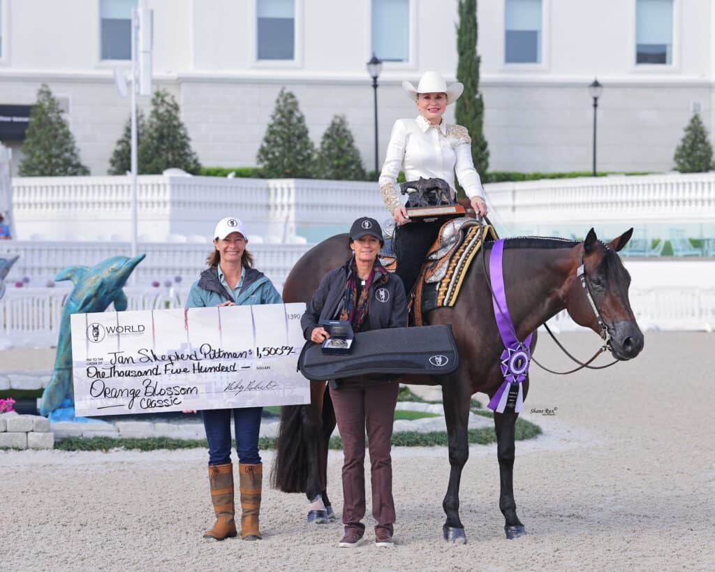 Three women pose at a horse show; one sits on a horse with award ribbons, while the others hold a large check and a case. The background shows a white fence and building.