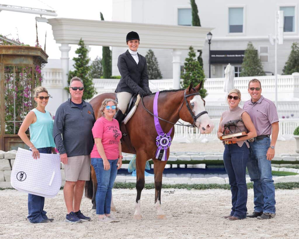 A rider sits on a horse adorned with ribbons, surrounded by five smiling people in casual attire, posing for a group photo at an equestrian event.
