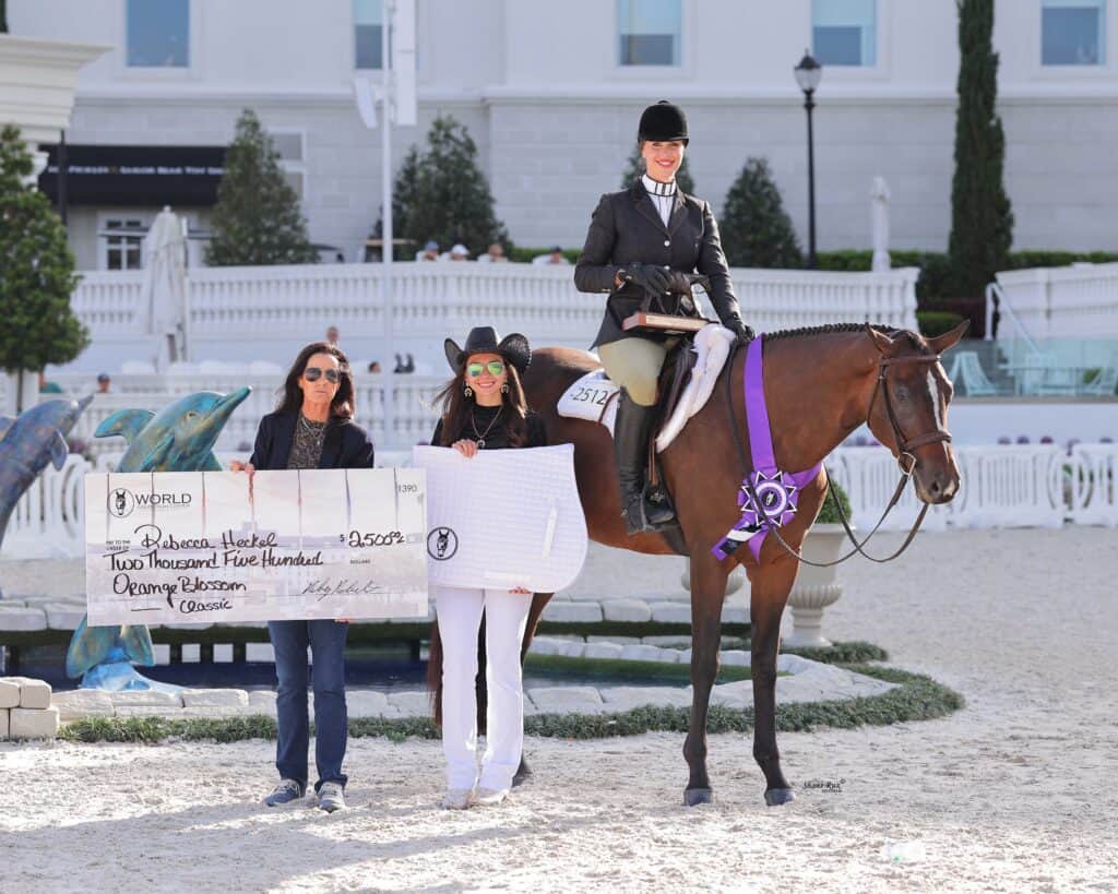 A woman on horseback wearing a ribbon poses with two people holding an oversized check for $2,500 at an outdoor equestrian event.