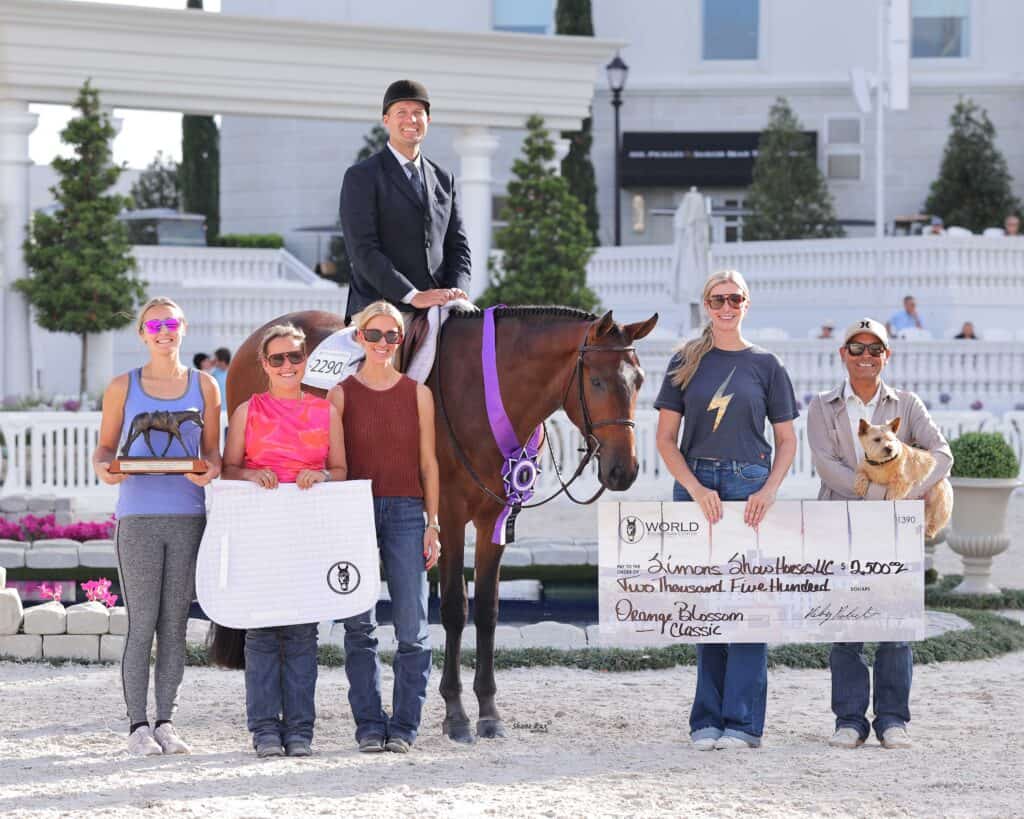 A group of five people and a dog pose with a horse, trophies, and a large check for $2,500 at an outdoor equestrian event.