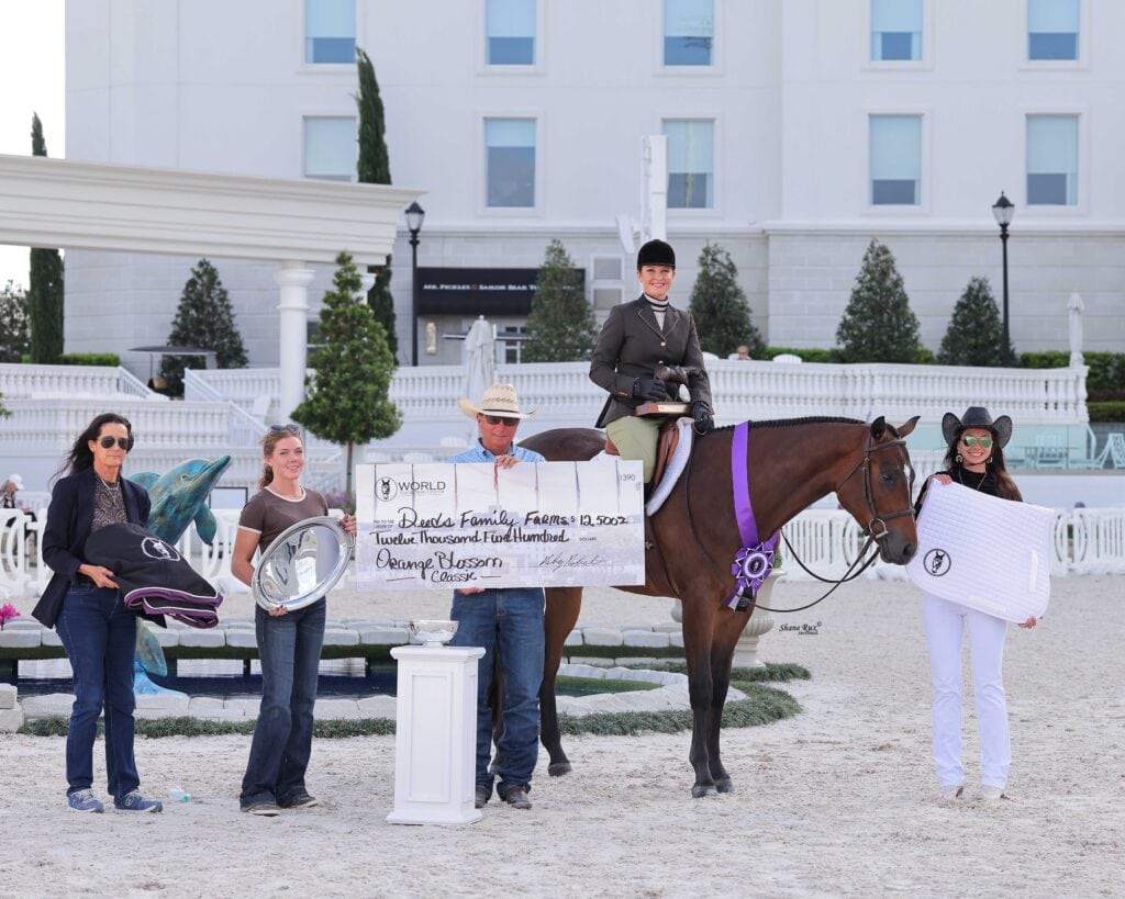 A group of people stands with a rider on a horse, holding a large check and various prizes at an outdoor equestrian event.