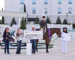 Five people and a horse pose outdoors; one person on horseback holds a ribbon, another holds a large check, while others display various prizes including a saddle, helmet, and blanket.