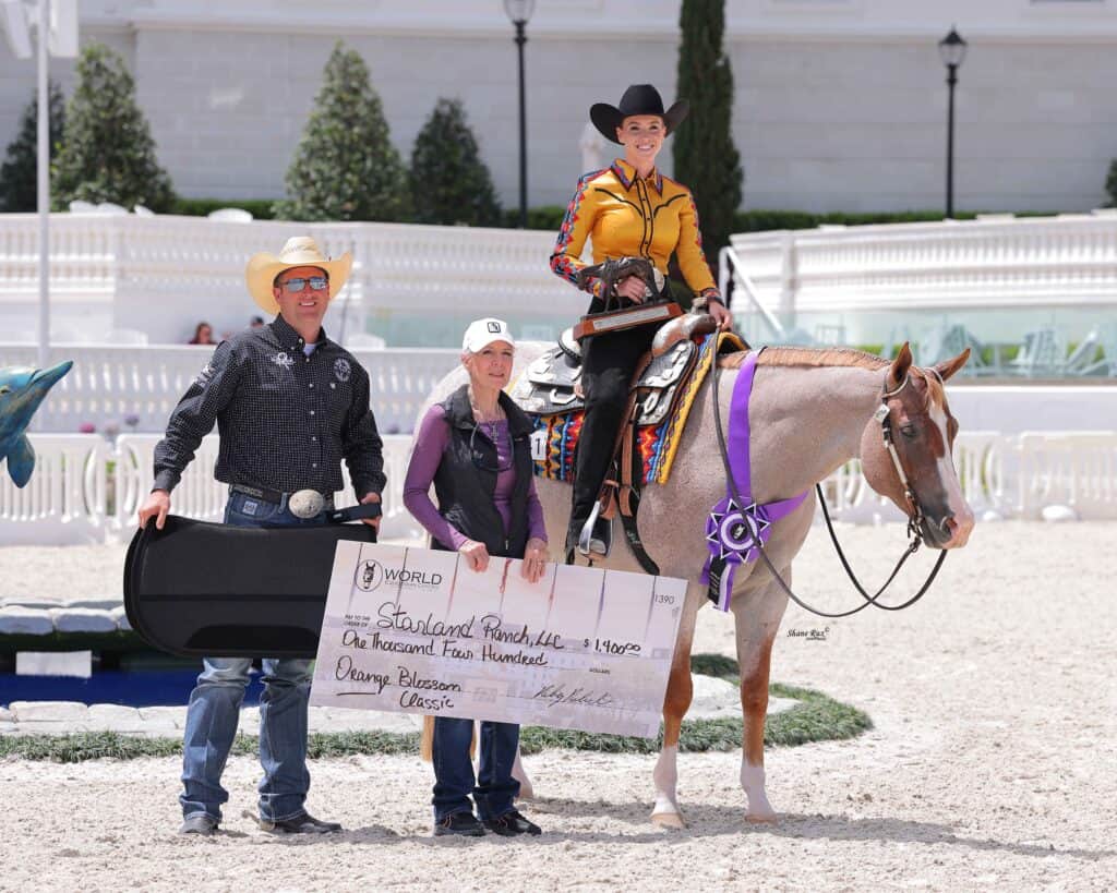 Three people pose with a horse; one is holding a large check and another is mounted on the horse wearing show attire. They appear to be at an equestrian event.