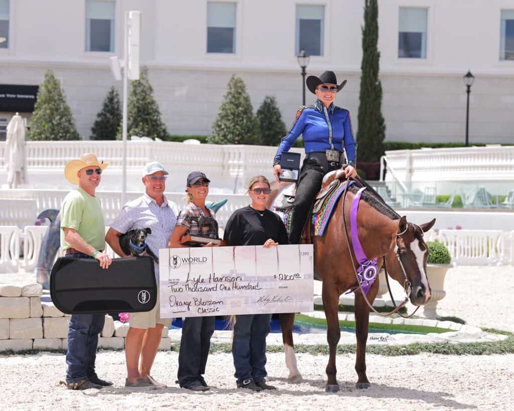 Five people and a horse pose outdoors; one woman on horseback holds a trophy ribbon. The group holds an oversized check labeled "Two Thousand One Hundred" and a guitar case.