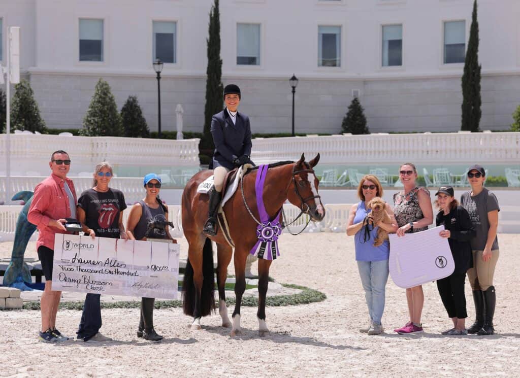 A group of people stand around a woman on a horse holding a purple ribbon; one person holds a large check, another holds a small dog.