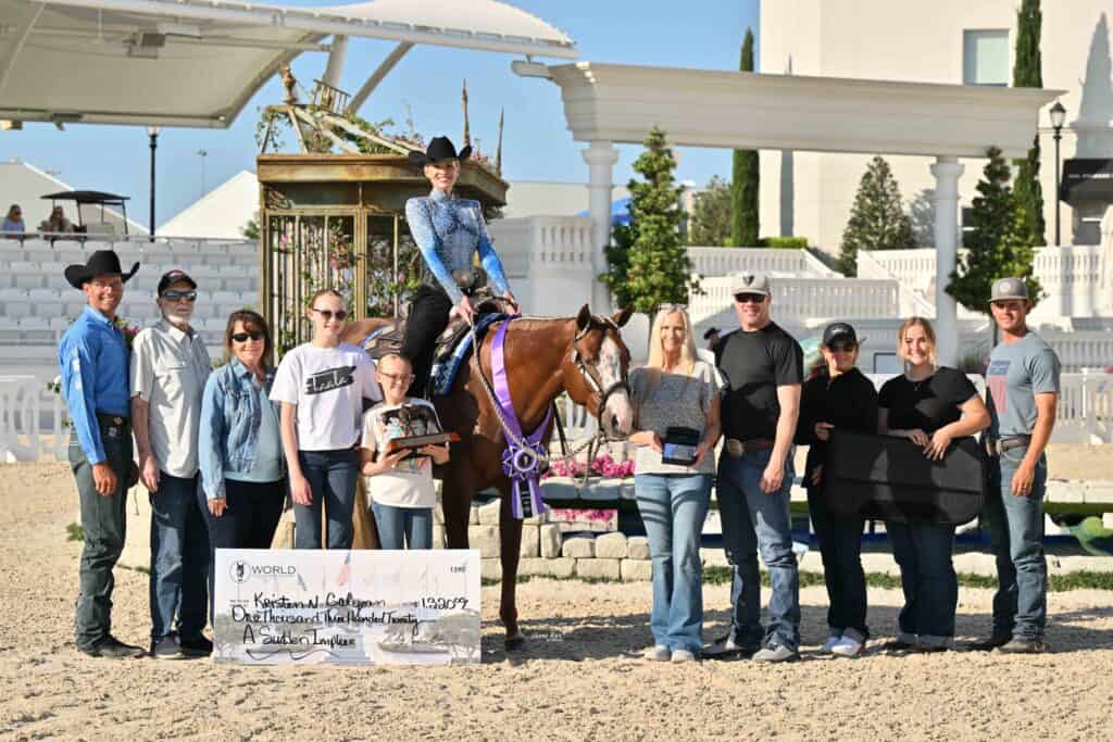 A group of people pose outdoors with a horse, a rider holding ribbons, a large winner’s check, and several awards.