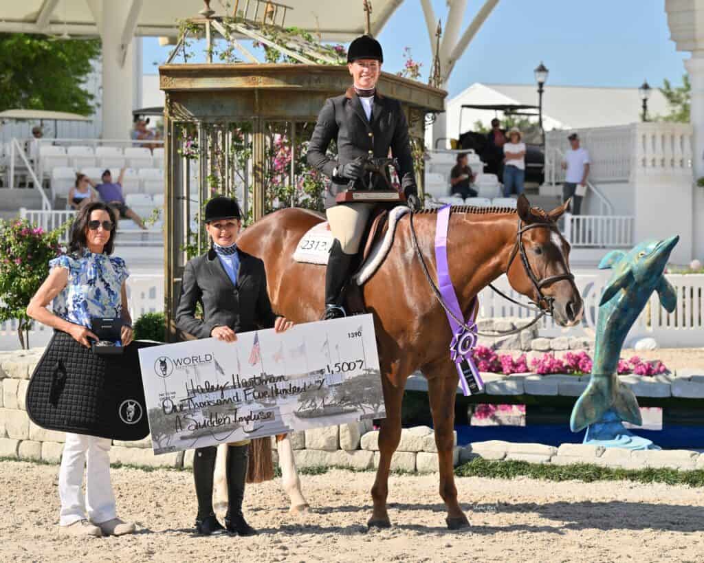 Three people pose with a horse at an equestrian event, holding a large check and a ribbon; an audience is seated in the background.