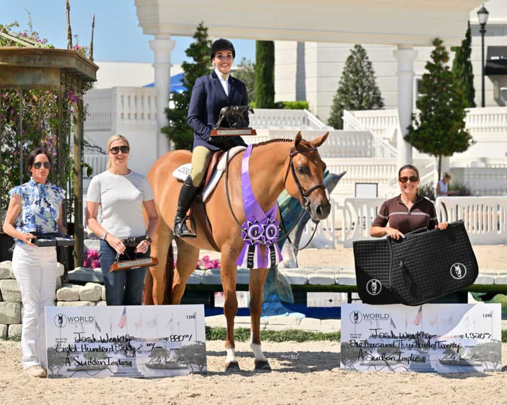 A rider on a horse poses with three women holding awards and oversized checks at an outdoor equestrian event.