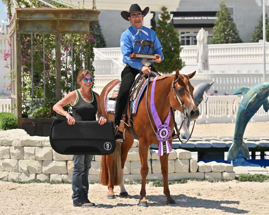 A man in a cowboy hat sits on a horse with a purple ribbon while holding a trophy. A woman stands beside him holding a black case. They are outdoors with statues and a white building in the background.