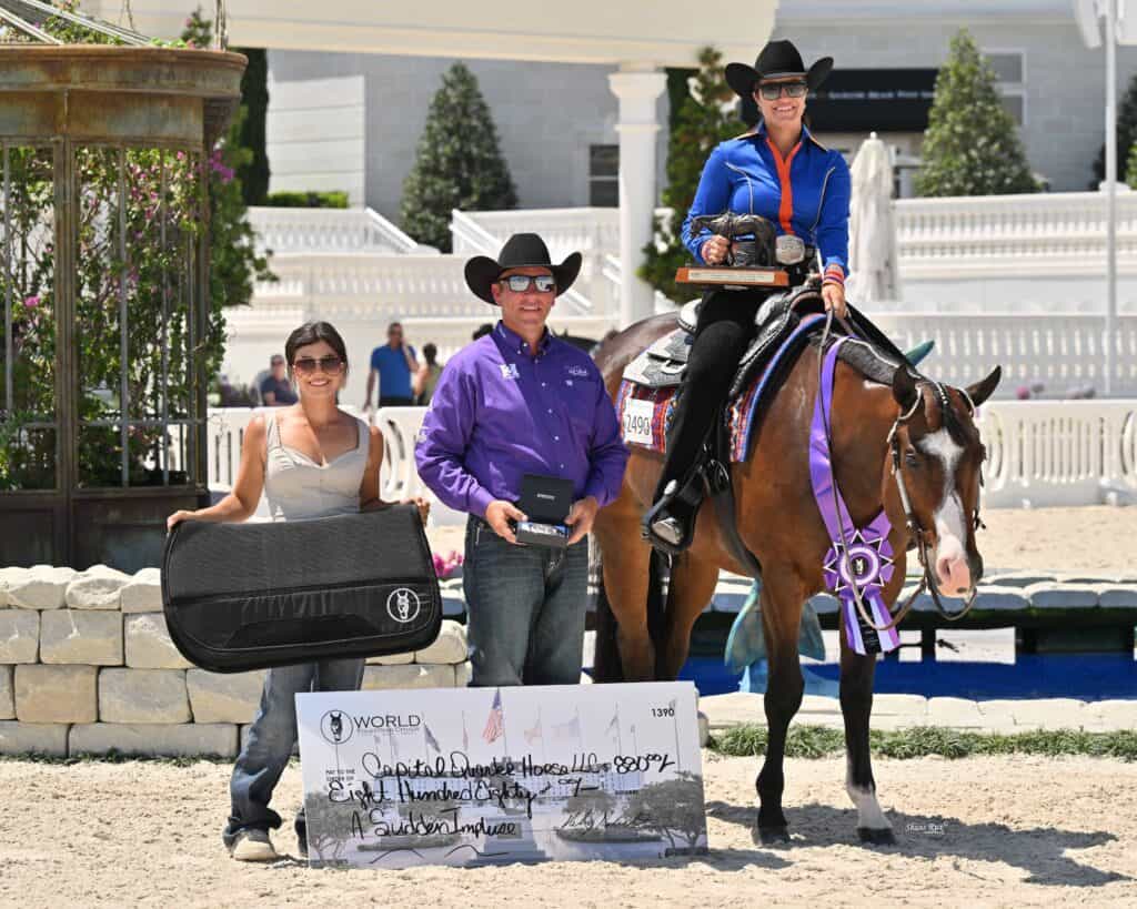 Three people pose with a horse, a large prize check, a black bag, and a medal at an outdoor equestrian event. The person on the horse holds a trophy and wears a competition outfit.
