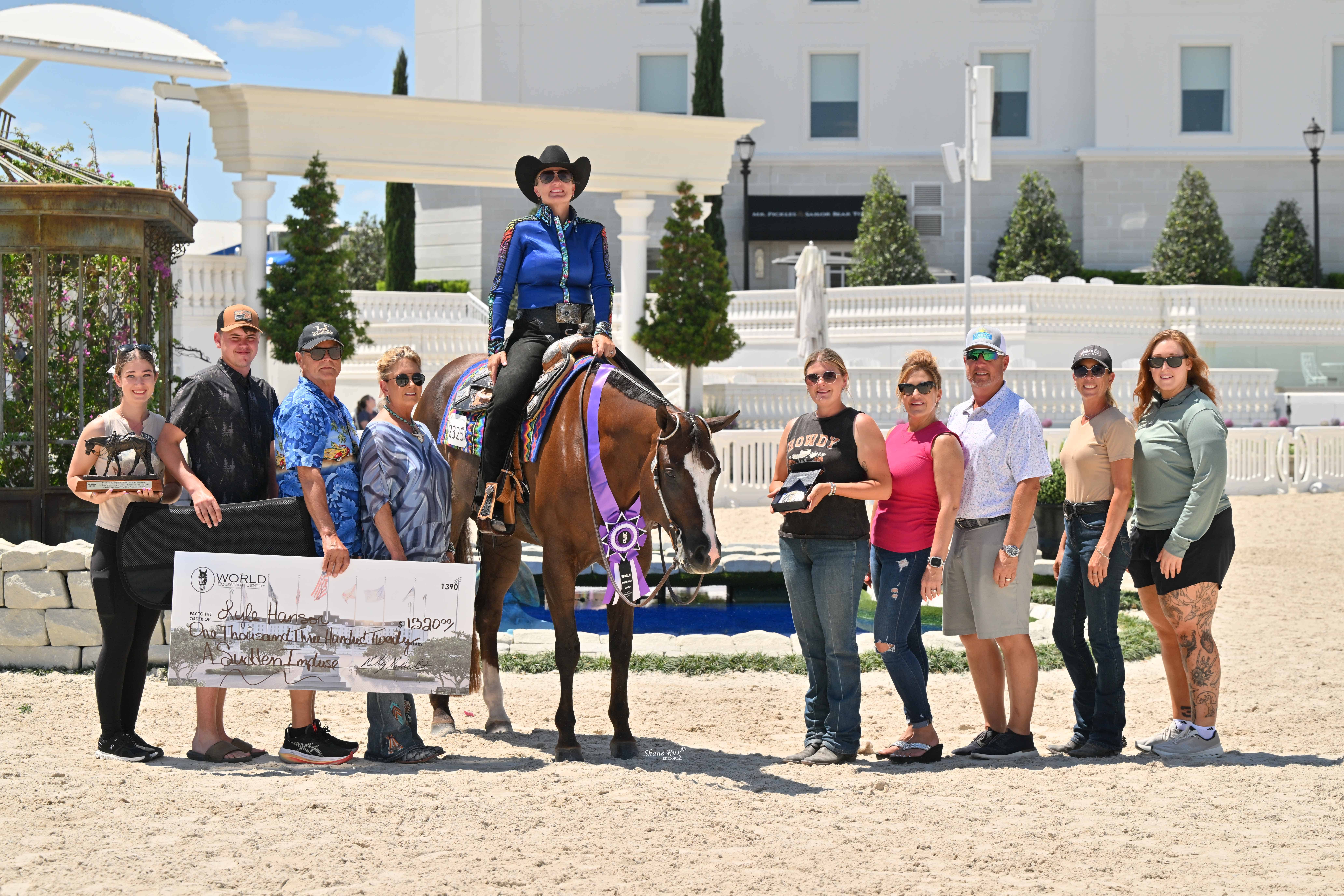A group of people pose with a woman on a horse wearing a ribbon, holding a trophy and an oversized check at an outdoor equestrian event.