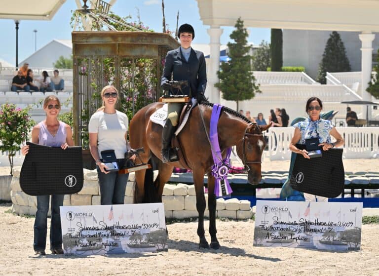 Four women at an equestrian event; one on horseback wearing riding attire with a ribbon, and three holding oversized checks and awards in an outdoor arena setting.