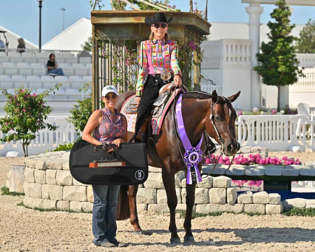 A woman on a horse with a prize ribbon sits next to another woman holding a trophy and a large black case; they pose in an outdoor equestrian arena.