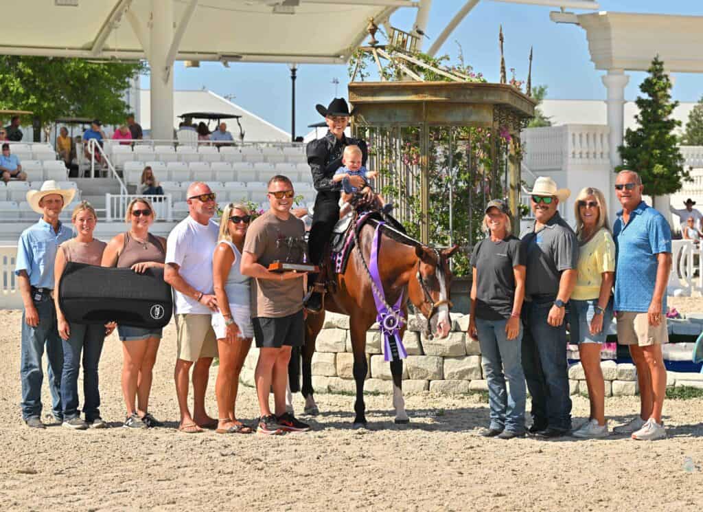 A group of people poses outdoors with a horse and rider holding a child. The rider and horse wear show attire and ribbons; the group stands on sandy ground with white seating in the background.