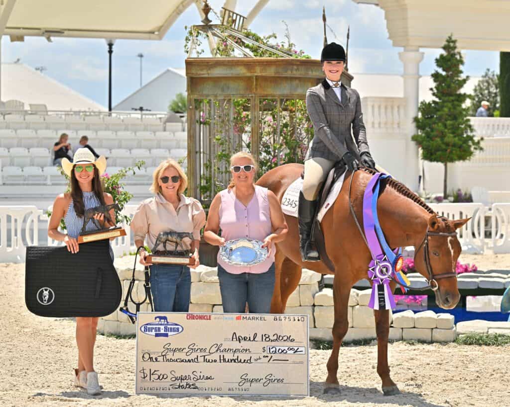 Four women and a horse in a show arena, with one woman on horseback and three holding prizes and a large check.