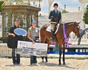 Three people pose with a horse; one person holds a silver tray, another holds a large prize check, and a rider in equestrian attire sits on the horse wearing a purple ribbon.