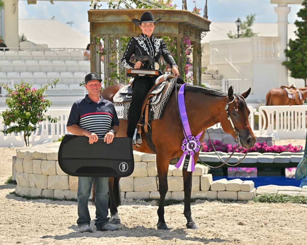 A person in Western show attire sits on a horse with a large purple and white ribbon. Another person stands beside them holding a black saddle pad.