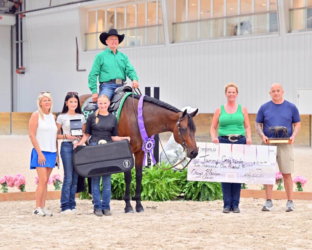 Seven people pose indoors with a horse, prize ribbons, trophies, and an oversized check. One person is seated on the horse, while others stand and hold awards.
