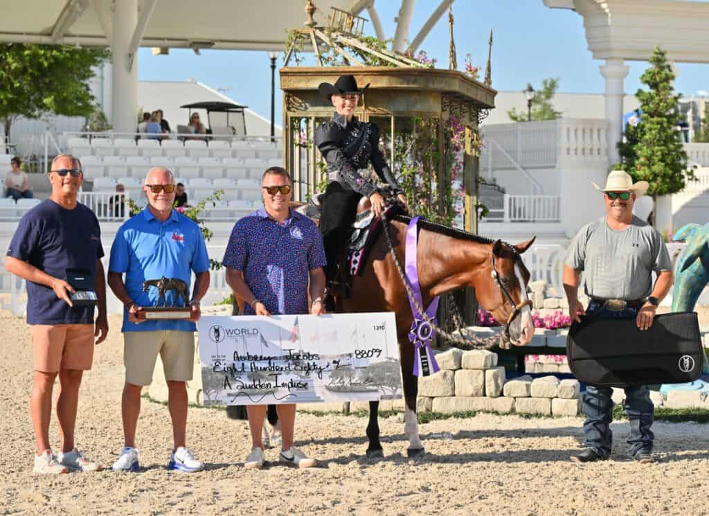 Five people and a horse pose outdoors with awards; one person on horseback holds a trophy, another holds a large presentation check displaying winnings.