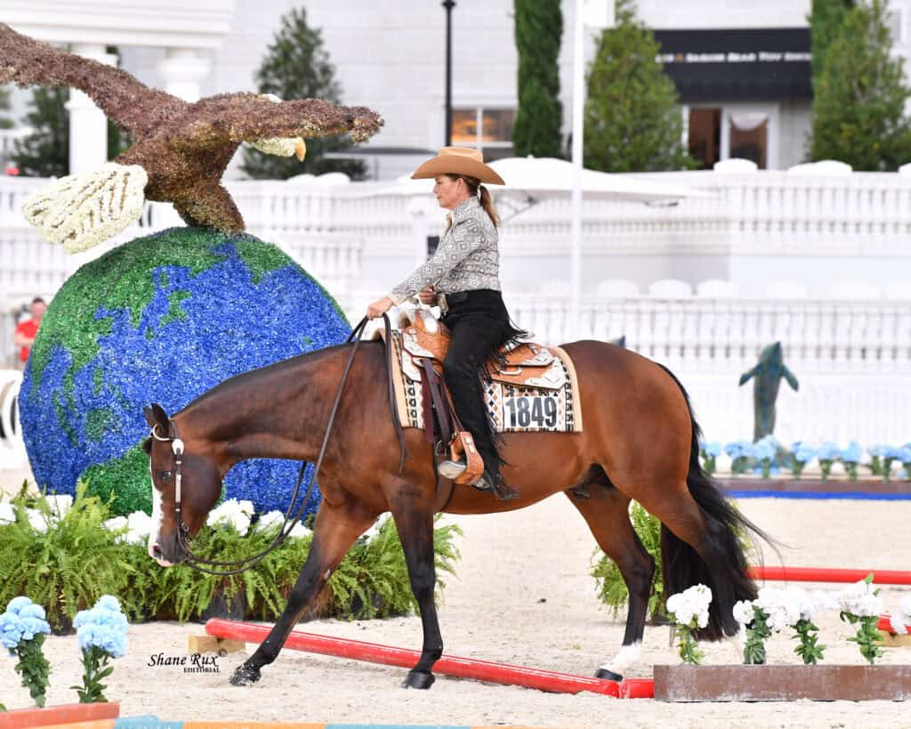 A woman in a hat rides a brown horse in an arena decorated with a large globe and eagle sculpture, surrounded by flowers and greenery.