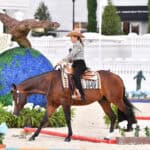 A woman in a hat rides a brown horse in an arena decorated with a large globe and eagle sculpture, surrounded by flowers and greenery.