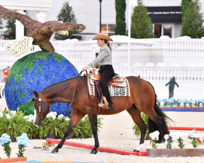 A woman in a hat rides a brown horse in an arena decorated with a large globe and eagle sculpture, surrounded by flowers and greenery.