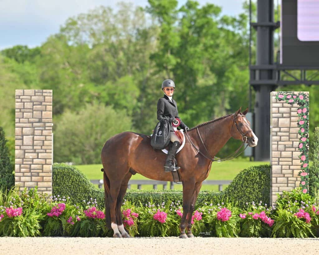 A rider in formal equestrian attire sits on a brown horse in an outdoor arena, surrounded by greenery, flowers, and decorative stone pillars.