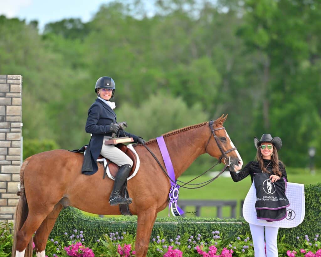 Equestrian rider on a brown horse with a purple ribbon, holding a trophy, while another woman stands beside them holding the horse’s lead.