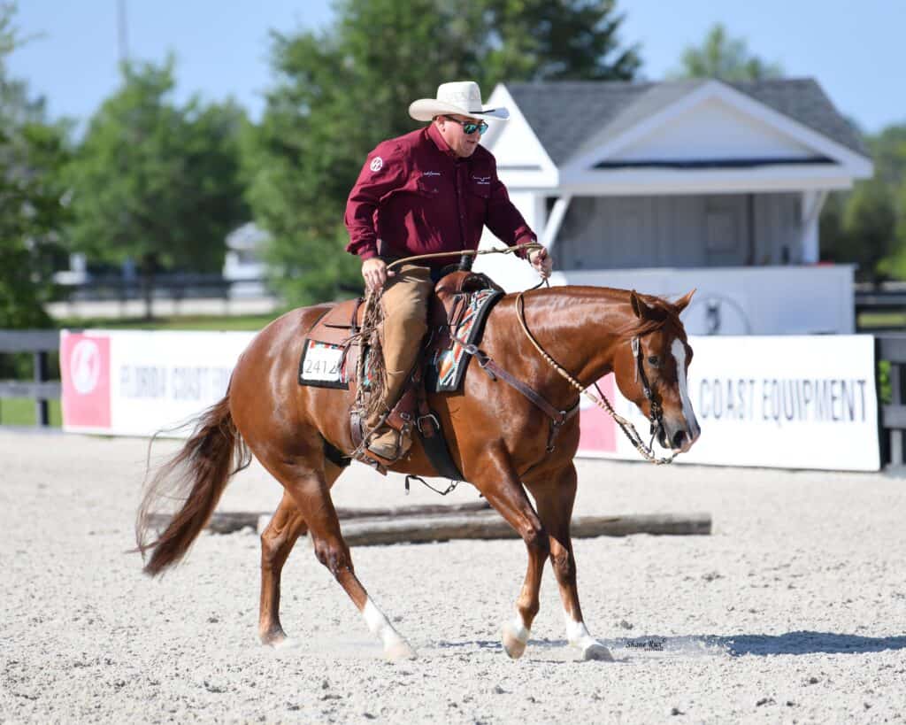 A person in a cowboy hat and maroon shirt rides a brown horse in an outdoor arena with a white building and trees in the background.