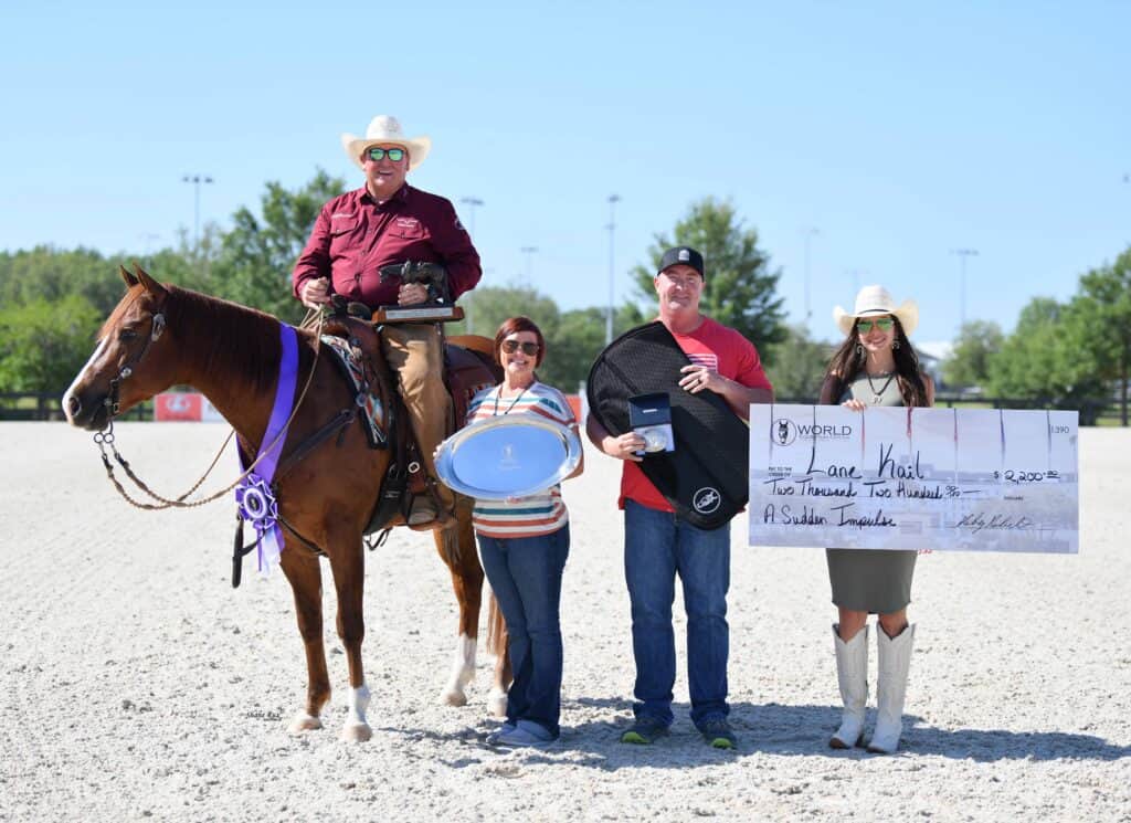 Four people pose outdoors with a horse and award items, including a large check, plate, and ribbon, at a competitive event.